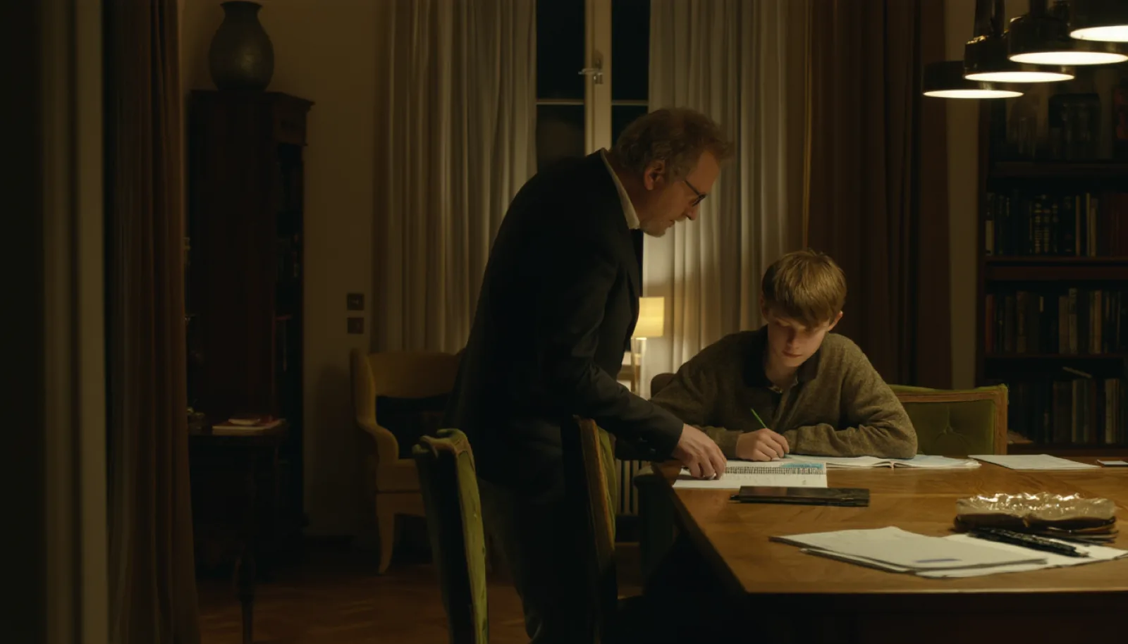 A tutor and a pupil working over an open exercise book at a dining table in evening light
